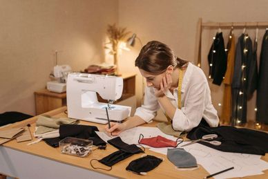 Woman designing face masks at a sewing table with sketches and fabric.