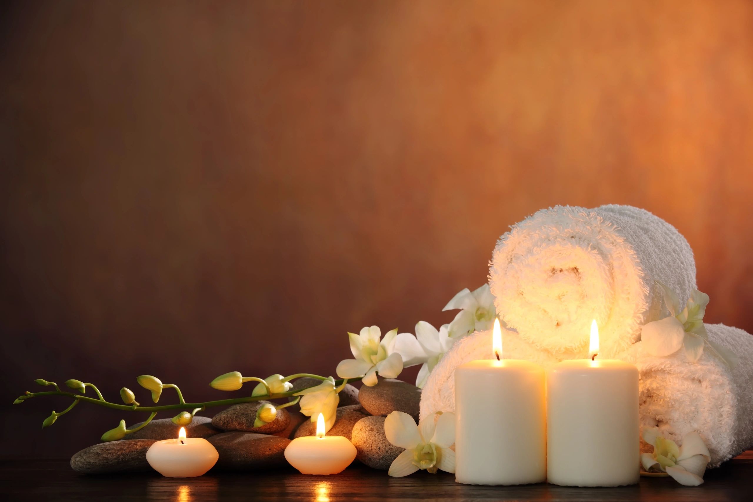 Towels, hot stones, lit candles, and white flower setting the tranquil ambiance for a massage.