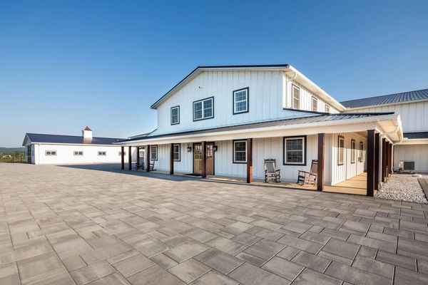 Modern white farmhouse with rocking chairs on a spacious paved porch under a clear blue sky.