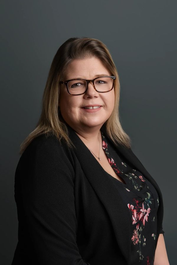 Smiling woman with glasses in a black floral top and blazer on a gray background.