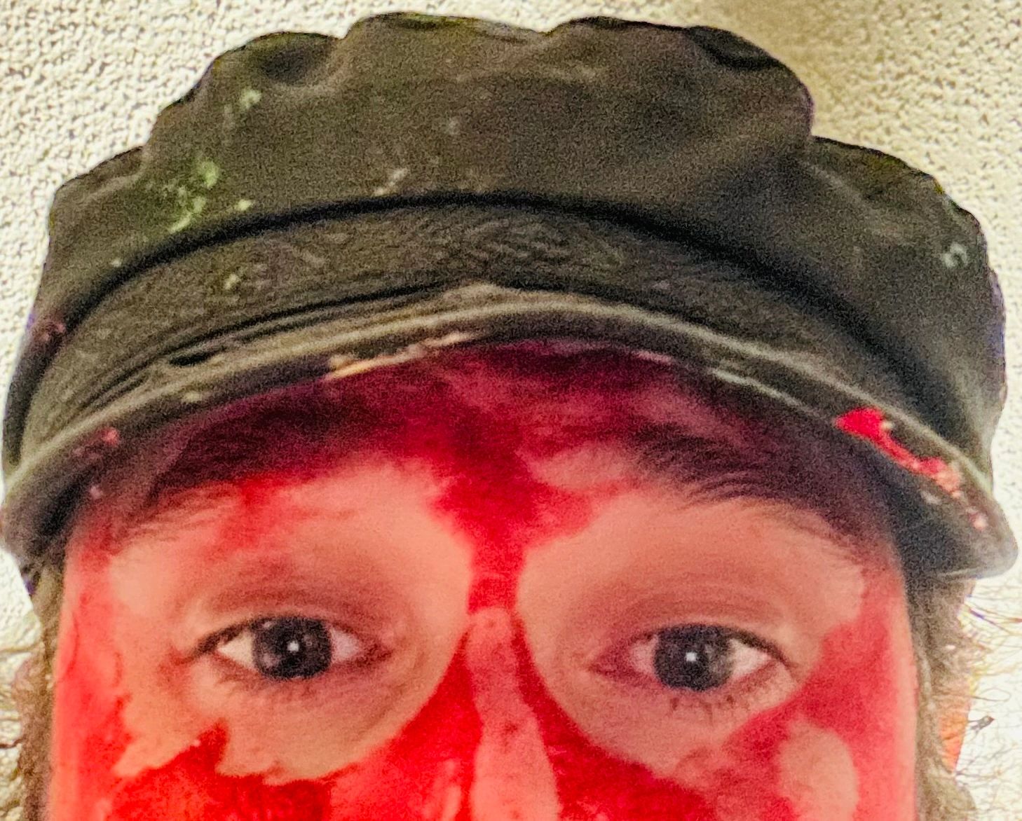 Close-up of a person with red paint on their face and wearing a green cap.
