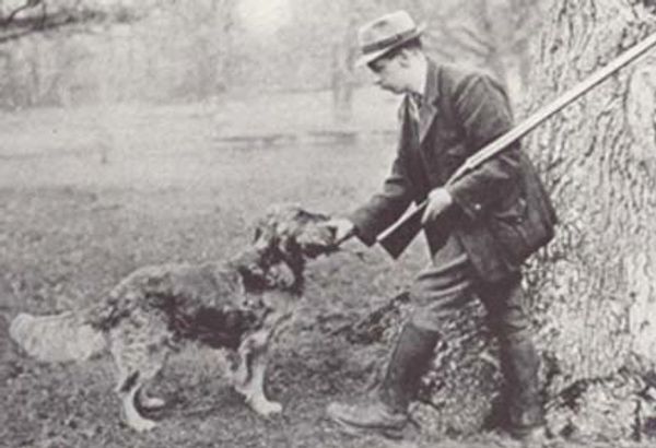 A man in vintage attire feeds a dog while holding a rifle.