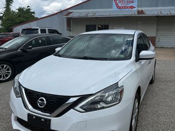 White Nissan sedan parked in front of a building under a partly cloudy sky.
