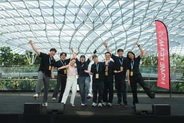 Group of eight people joyfully posing on stage under a glass roof.