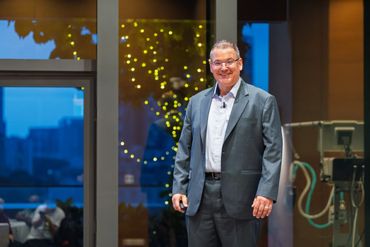 Smiling man in a suit standing indoors with festive lights in the background.