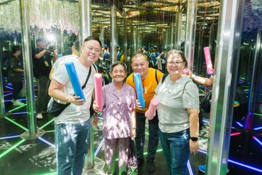 Four smiling people holding colorful foam sticks in a mirrored room with neon floor lights.