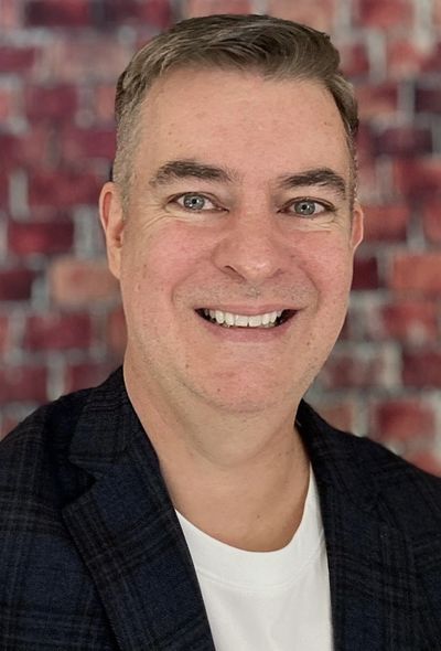 Smiling man in a dark checkered blazer with a blurred brick wall background.