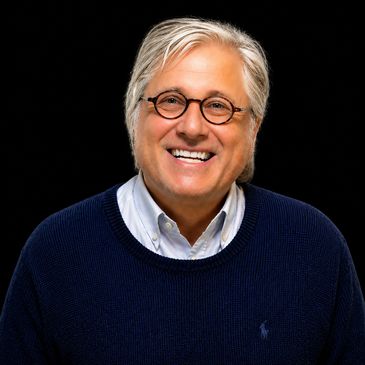 Smiling man with round glasses and white hair in a navy sweater.