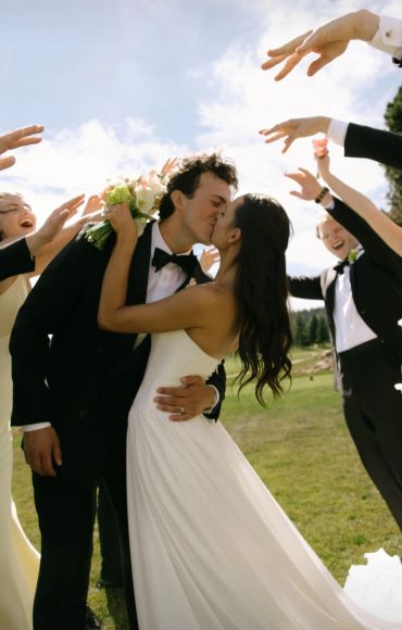 Bride and groom sharing a kiss outdoors with friends applauding.
