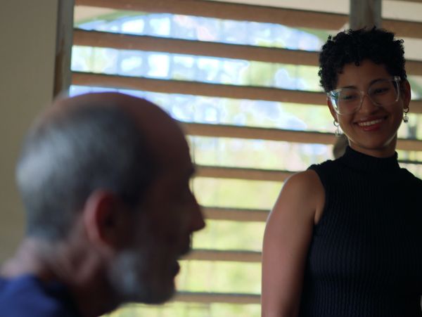 A smiling woman listens to an older man speaking indoors near a window.
