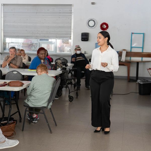 A woman speaks to a group of elderly people in a community room.