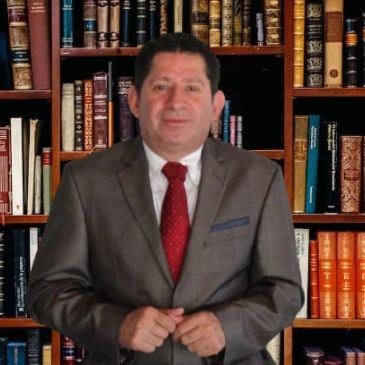 Hombre de traje marrón y corbata roja frente a estantería con libros.