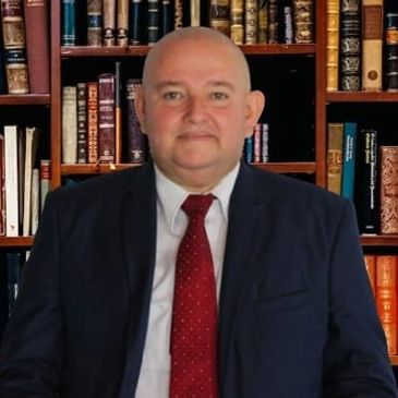 Hombre calvo con traje y corbata roja frente a estantería de libros.