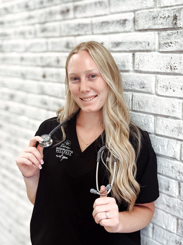 Smiling healthcare professional with long blonde hair and a stethoscope against a white brick wall.