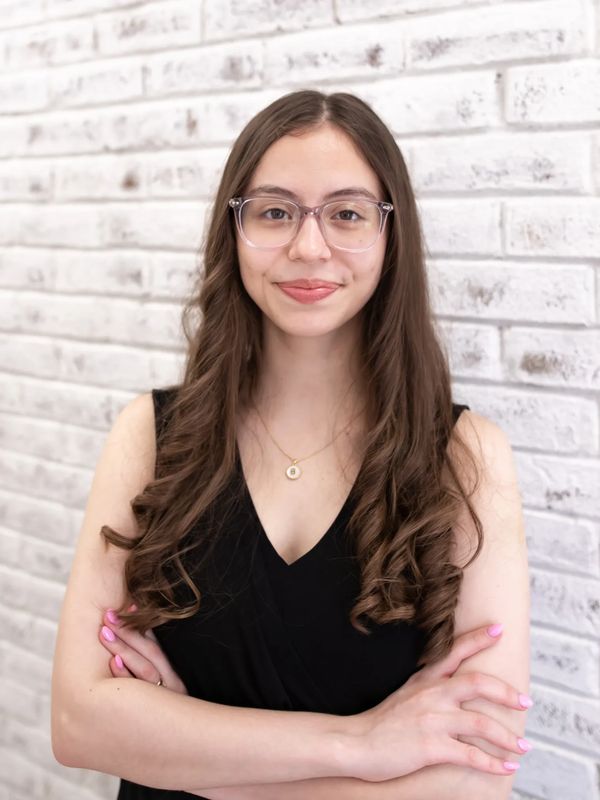 Young woman with glasses and long curly hair smiles confidently with arms crossed.
