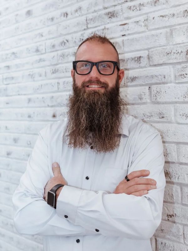 Confident man with a long beard and glasses stands against a white brick wall.