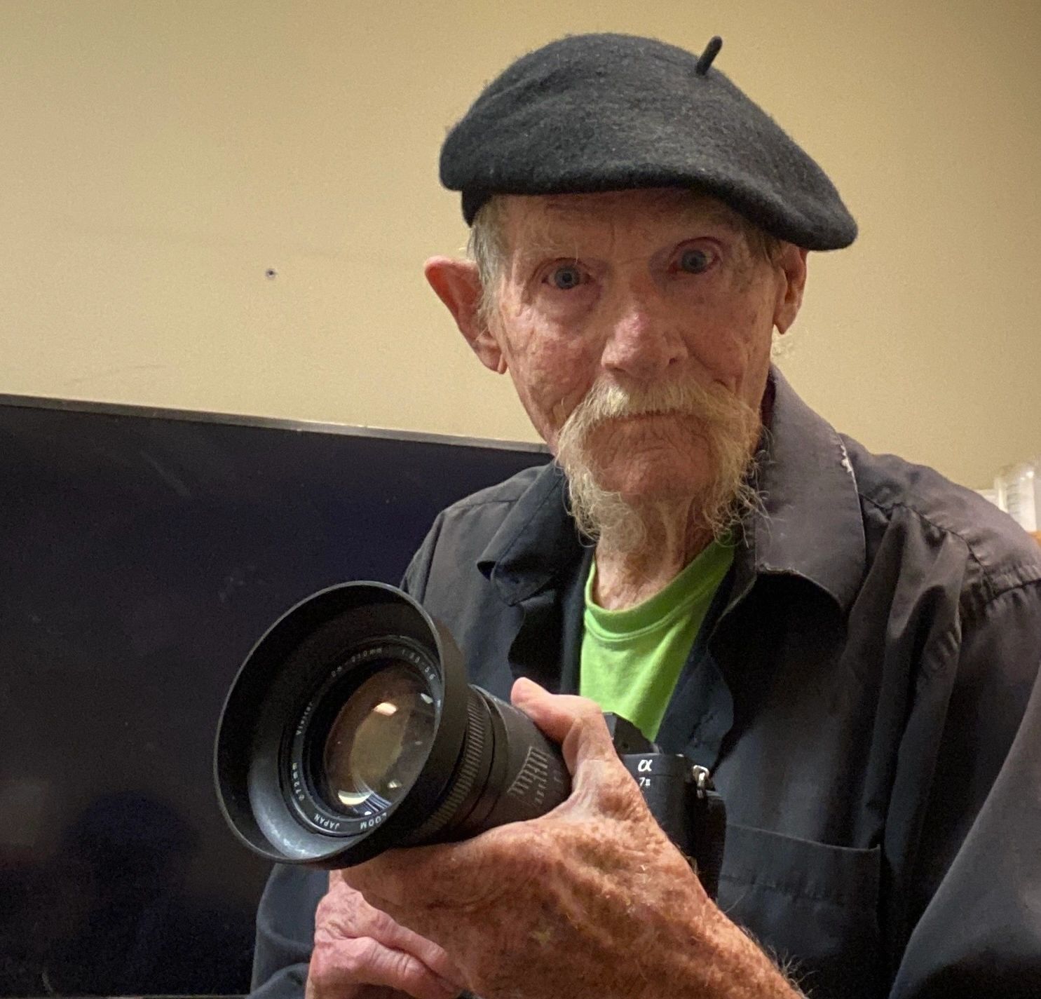 Elderly man in a black beret holding a camera with a large lens.