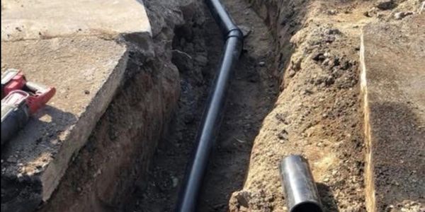 A black pipeline being installed in a dug trench beside a concrete surface.