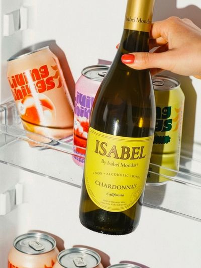 Hand holding a bottle of Isabel non-alcoholic Chardonnay wine in a fridge with colorful Living Things cans.