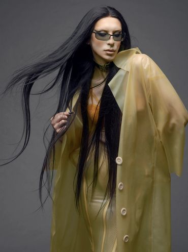 Fashion model in a translucent yellow coat and sunglasses with flowing black hair.
