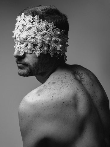 Black and white photo of a man with lace covering his eyes, showing his back and tattoo.