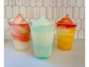 Three colorful frozen slush drinks in clear plastic cups on a white surface.