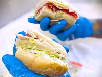 Two sandwich halves held with blue gloves, filled with lettuce, tomato, and turkey.