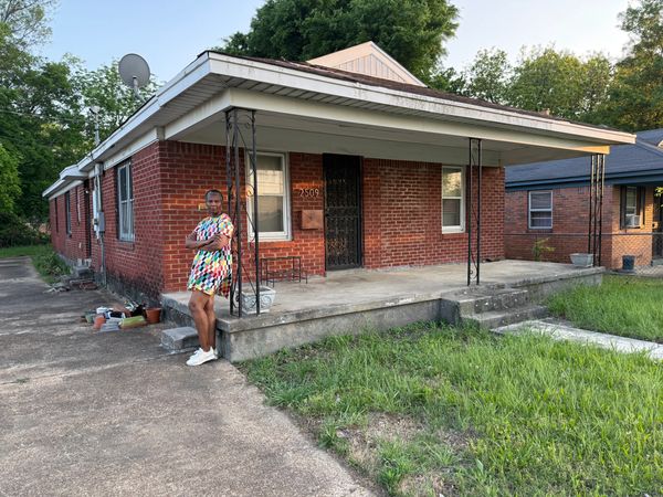 Woman in colorful dress standing outside a brick house porch.