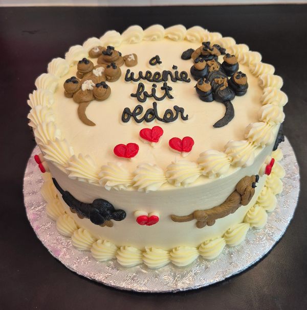 White cake with dachshund dog decorations and 'a weenie bit older' text.