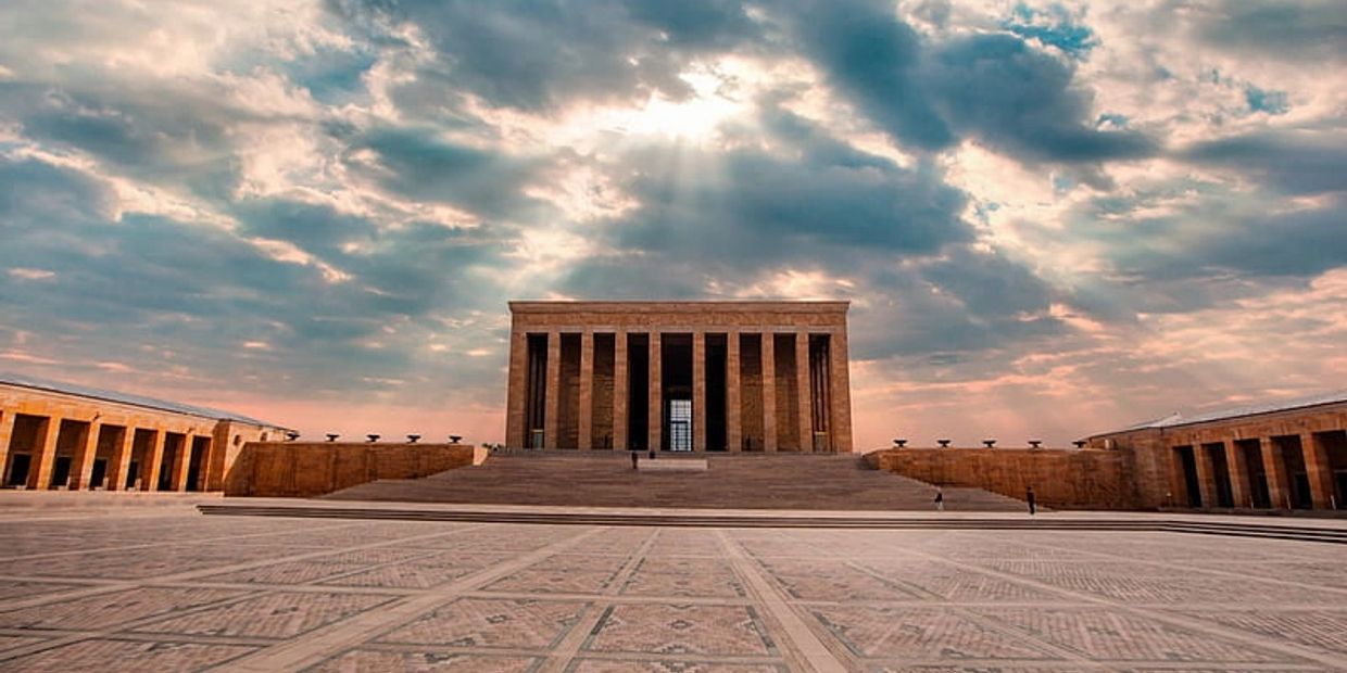 Sun rays break through clouds over a grand, columned monument with wide steps and patterned ground.