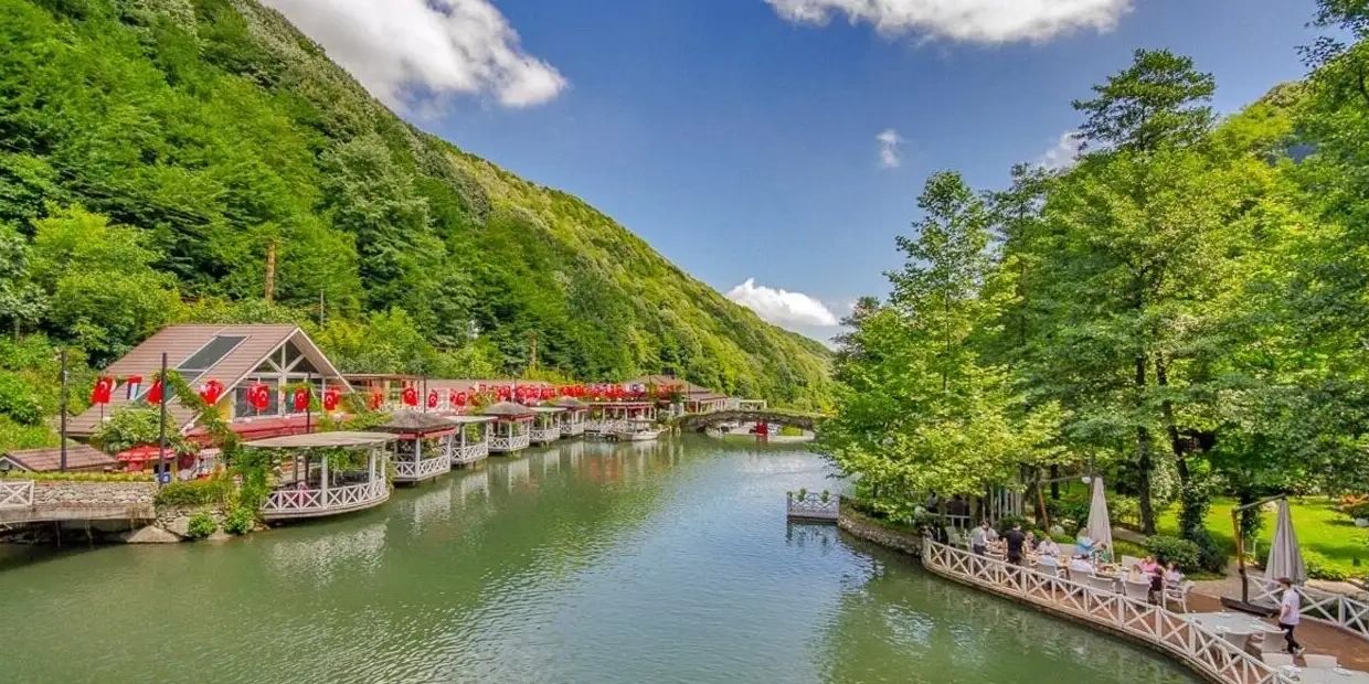 A peaceful riverside scene with green hills and Turkish flags lining the walkway.