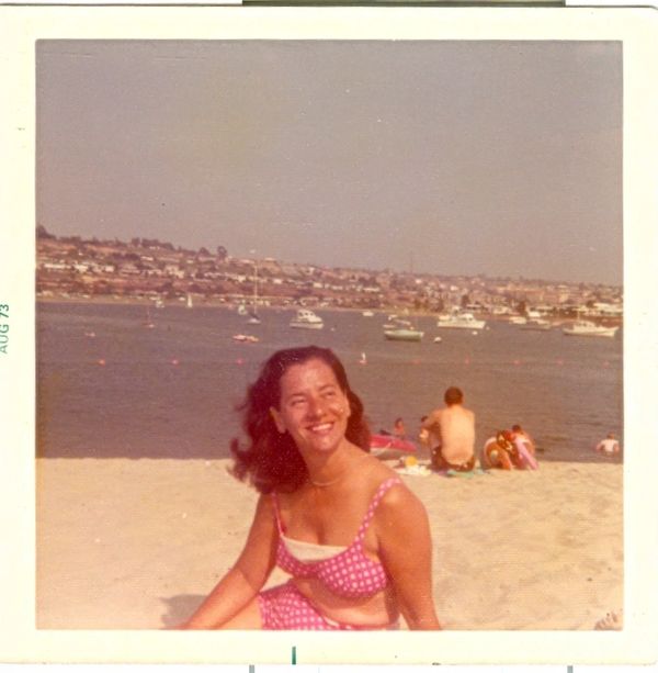 Smiling woman in a pink polka dot swimsuit at a sunny beach with boats in the background.