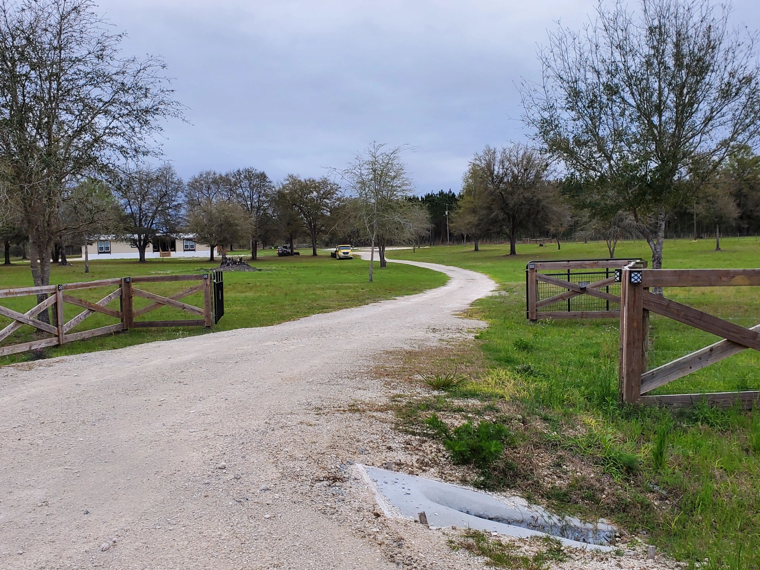Gravel driveway leading to a house with open wooden gates and scattered trees.