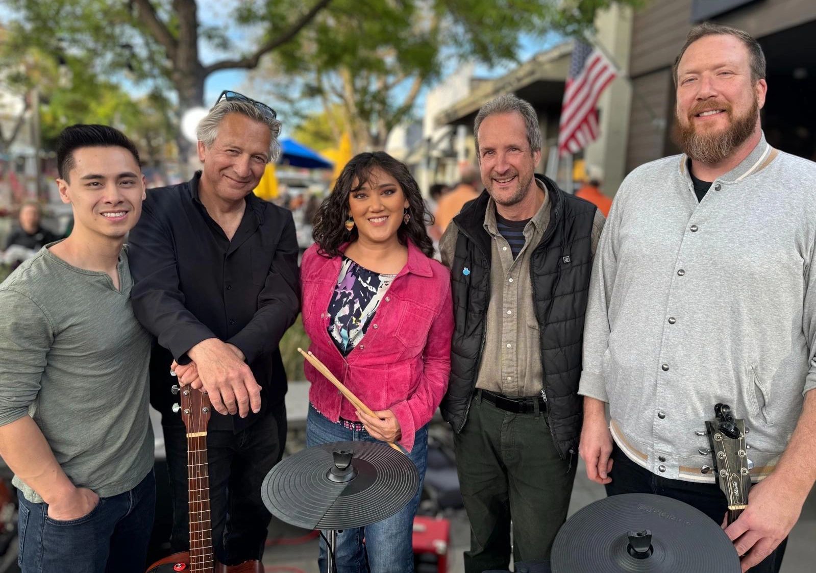 Five musicians posing outdoors with instruments, smiling warmly.