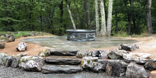 Stone fire pit with natural rock seating area in a forest setting.