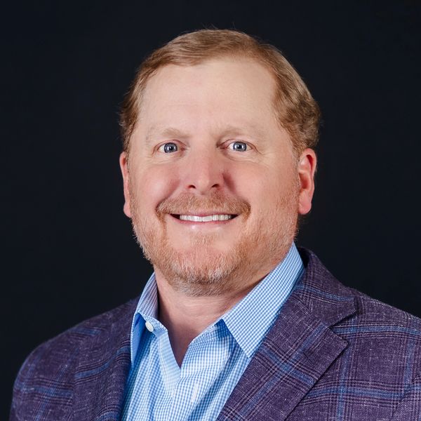 Smiling man in a blue checkered shirt and purple blazer against a black background.