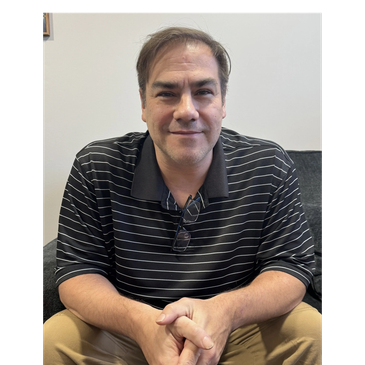 Man sitting on a couch, wearing a black striped polo and khaki pants.