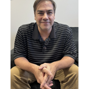 Man sitting on a couch, wearing a black striped polo and khaki pants.