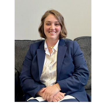 A smiling woman in a navy blue suit sitting on a gray couch.