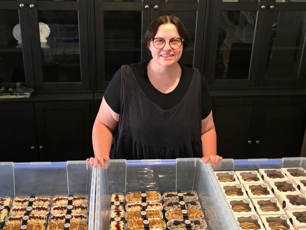 A smiling woman stands behind trays of assorted pastries.