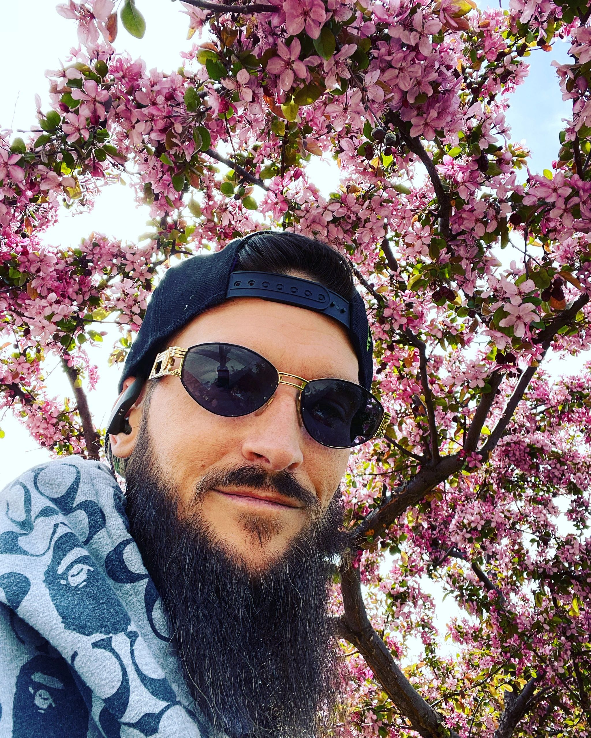 Man with sunglasses and a cap under a blooming pink tree.