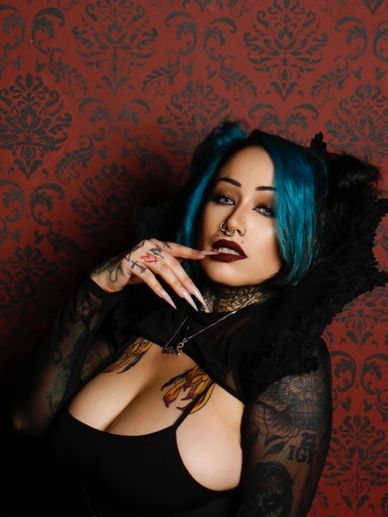Woman with blue hair and tattoos poses against a patterned wall.
