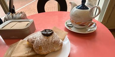 Croissant with chocolate and tea setup on a pink table by a window.