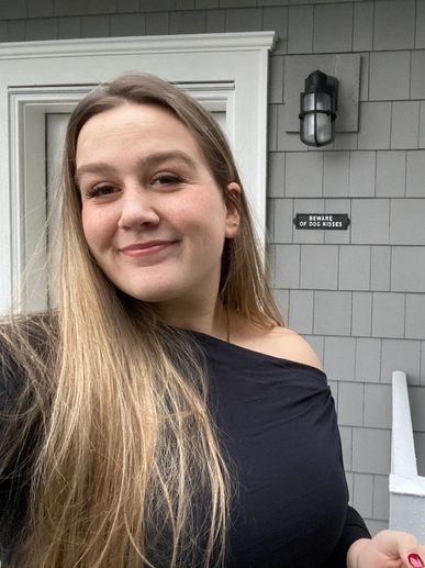 Smiling woman with long blonde hair standing outside near a door and a humorous sign.
