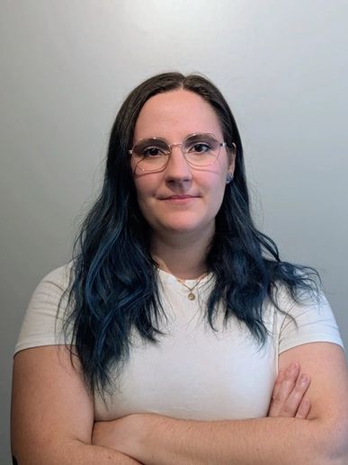 Confident woman with blue hair and glasses stands with arms crossed.