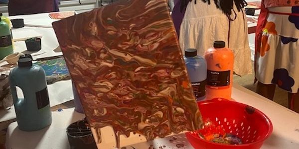 Person holding a canvas with a marbled paint pattern over a table with paint bottles.