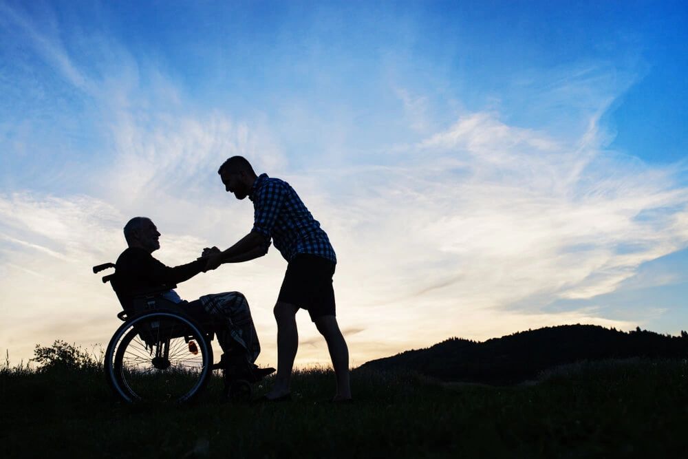 A silhouette of a support person holding a man’s hand that is sitting in a wheelchair.