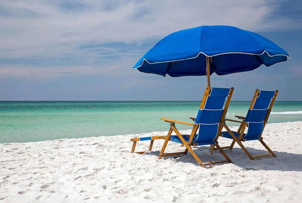 Two blue beach chairs and umbrella on white sand by turquoise ocean.