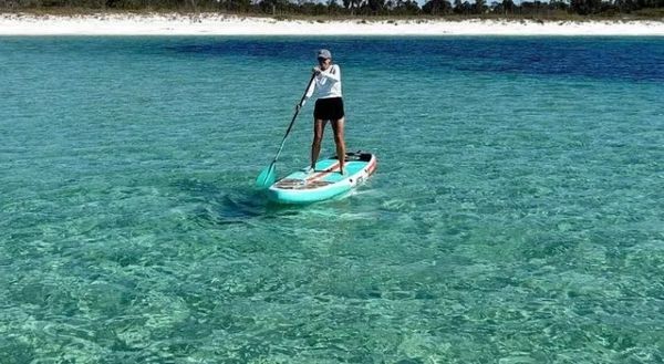 Person paddleboarding on clear turquoise water near a sandy shore.