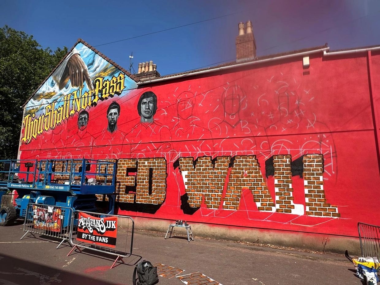 Mural in progress on a red wall with portraits and bold text.